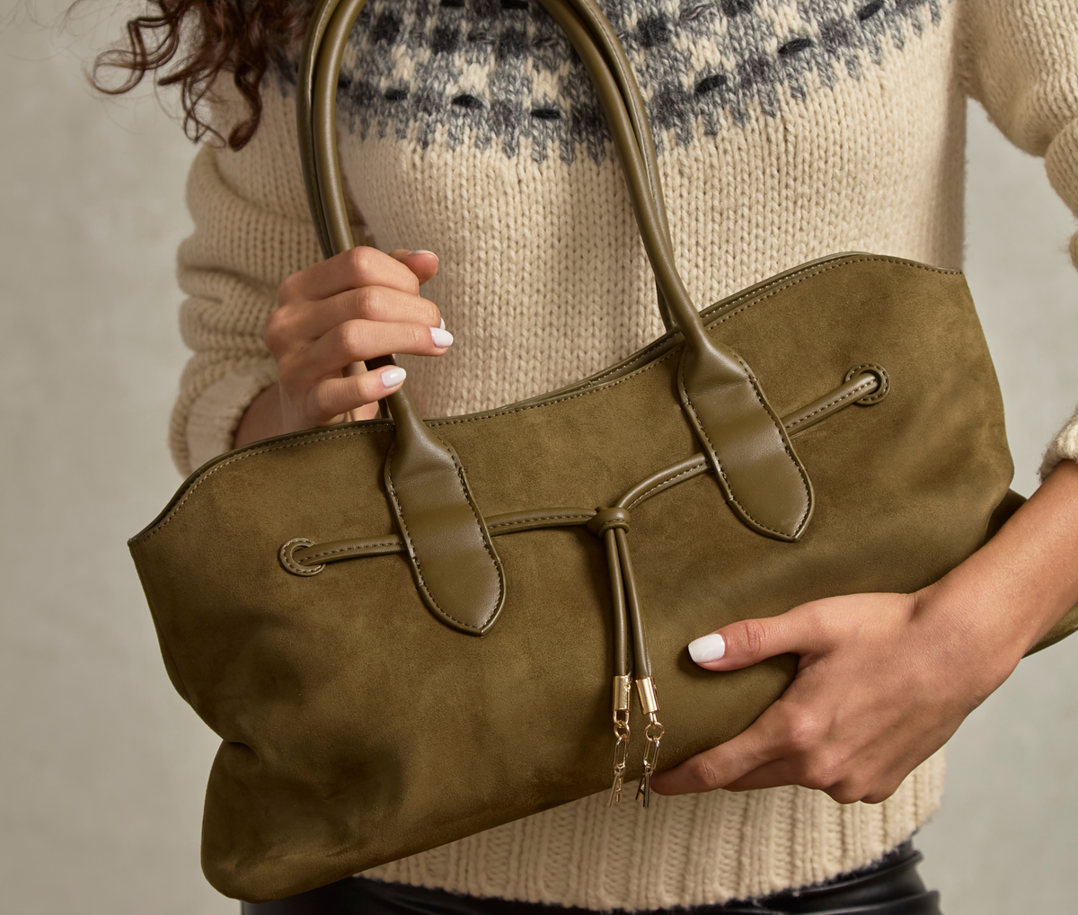 An olive green suede tote bag with smooth leather trim and dual top handles, held by a person wearing a cream knit sweater.