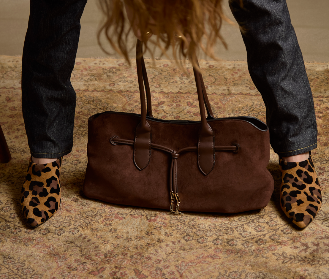 A brown suede duffel bag with dark brown leather handles and trim, shown on a patterned rug with a person's legs in dark jeans and leopard print shoes visible around it.