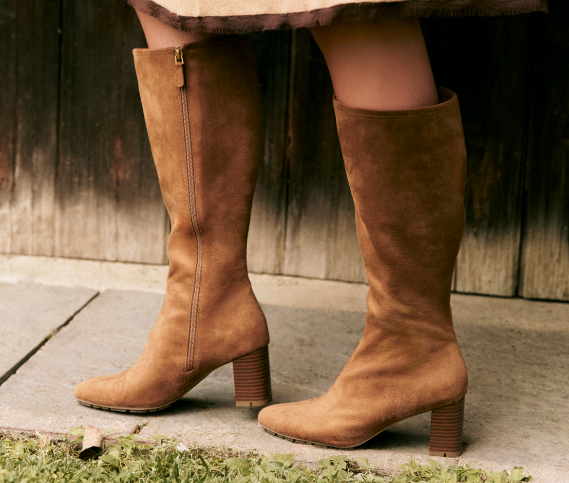 A close-up of a person wearing knee-high brown suede boots with block heels, standing on a stone surface against a weathered wooden wall.