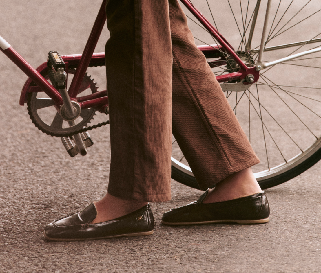 A person wearing brown wide-leg trousers and black leather loafers stands beside a red bicycle on a paved surface.