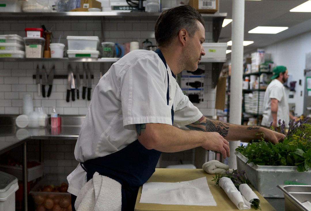 A male chef in a white uniform and navy apron works in a commercial kitchen, reaching toward fresh green herbs on a counter. Another person is visible in the background.
