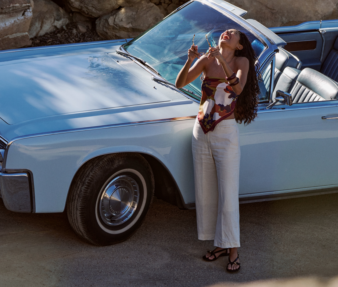 Woman standing next to a vintage car, holding a drink, with a scenic background.