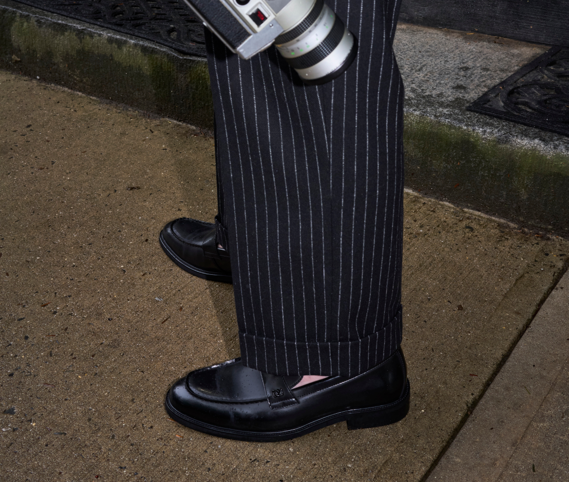 A close-up of a person's legs wearing black pinstripe trousers and black leather loafers, standing on a concrete sidewalk.