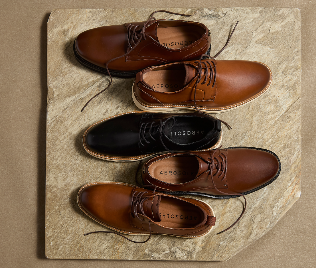 Four Aerosoles lace-up shoes arranged on a stone slab: two tan/cognac leather shoes, one black leather shoe, and one brown leather shoe. The shoes are photographed from above on a beige surface.